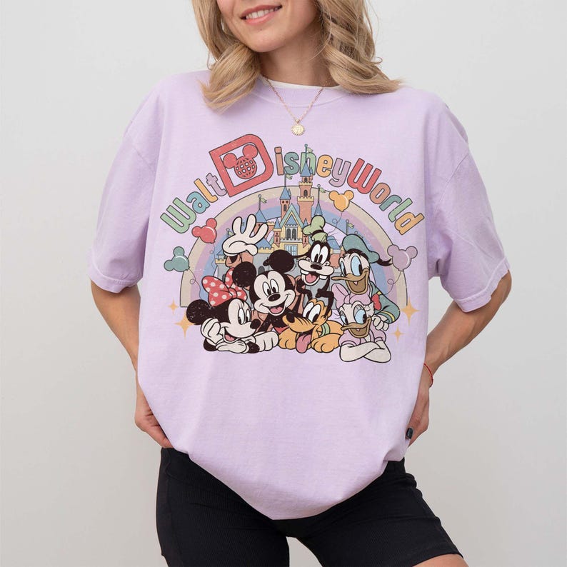 May include: A light purple t-shirt featuring a colorful graphic of Disney characters, including Mickey Mouse, Minnie Mouse, Donald Duck, Goofy, and Pluto, with the text "Walt Disney World" above them. The design includes a rainbow and a castle.