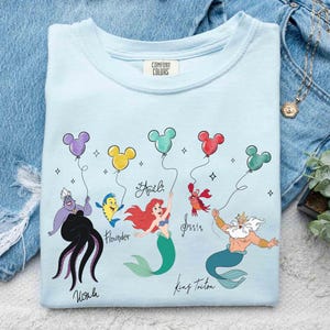 Puede incluir: Camiseta azul claro con un gr&aacute;fico colorido de personajes de La Sirenita, incluyendo a Ariel, Flounder y el Rey Trit&oacute;n. El dise&ntilde;o incluye globos y nombres de personajes. La camiseta est&aacute; hecha de un material suave.