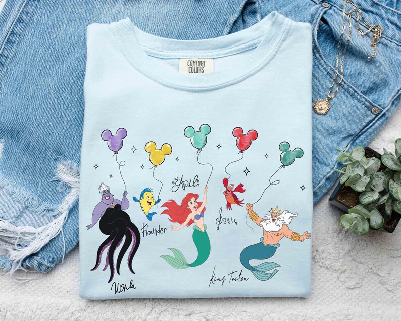 Puede incluir: Camiseta azul claro con un gr&aacute;fico colorido de personajes de La Sirenita, incluyendo a Ariel, Flounder y el Rey Trit&oacute;n. El dise&ntilde;o incluye globos y nombres de personajes. La camiseta est&aacute; hecha de un material suave.