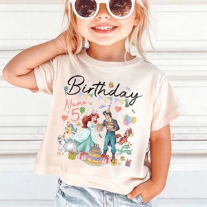 Pode incluir: Camiseta bege com um gr&aacute;fico colorido de anivers&aacute;rio. O design inclui a palavra "Birthday", o n&uacute;mero "5", personagens de desenhos animados, um bolo e presentes. Esta camiseta infantil &eacute; adequada para uma festa de anivers&aacute;rio.