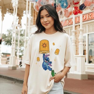 May include: A cream-colored short-sleeve t-shirt featuring a graphic print of a blue alien character hanging from a large yellow lantern with a sun symbol, accompanied by a small green creature and several smaller floating yellow lanterns.