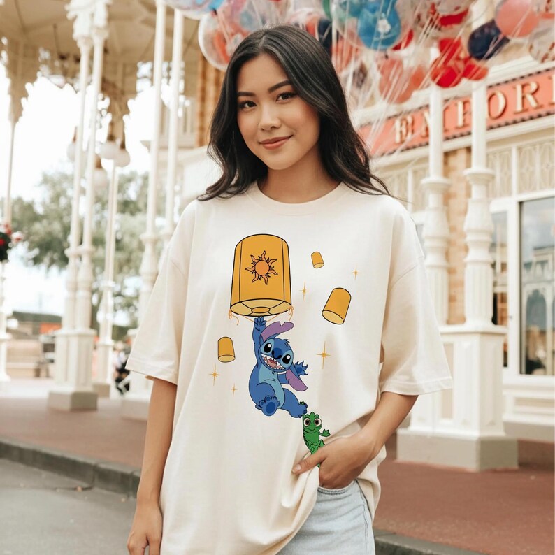 May include: A cream-colored short-sleeve t-shirt featuring a graphic print of a blue alien character hanging from a large yellow lantern with a sun symbol, accompanied by a small green creature and several smaller floating yellow lanterns.