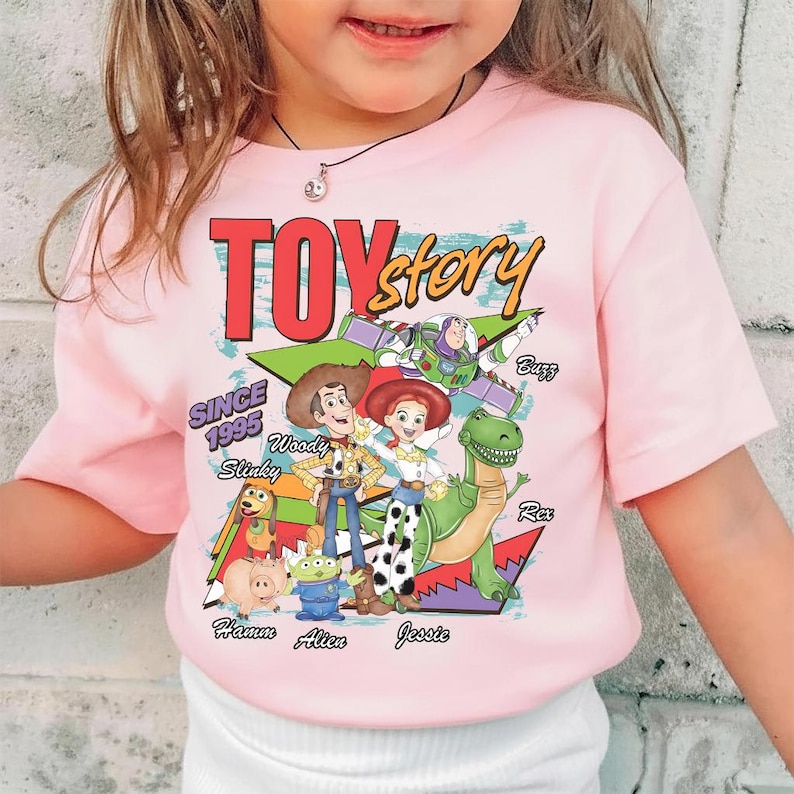 K&ouml;nnte beinhalten: Hellrosa T-Shirt mit den Worten "TOY STORY" in Rot und Gelb, sowie Illustrationen von Woody, Buzz, Jessie, Rex und anderen Charakteren. Das T-Shirt enth&auml;lt auch den Text "SINCE 1995".