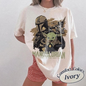 May include: Ivory-colored t-shirt featuring a graphic of The Mandalorian and Grogu. The design includes the text "MANDALORIAN - THIS IS THE WAY". The shirt is a Comfort Colors brand.