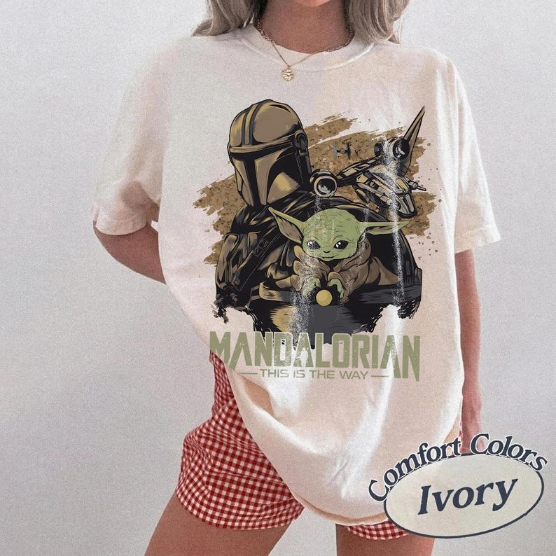 May include: Ivory-colored t-shirt featuring a graphic of The Mandalorian and Grogu. The design includes the text "MANDALORIAN - THIS IS THE WAY". The shirt is a Comfort Colors brand.