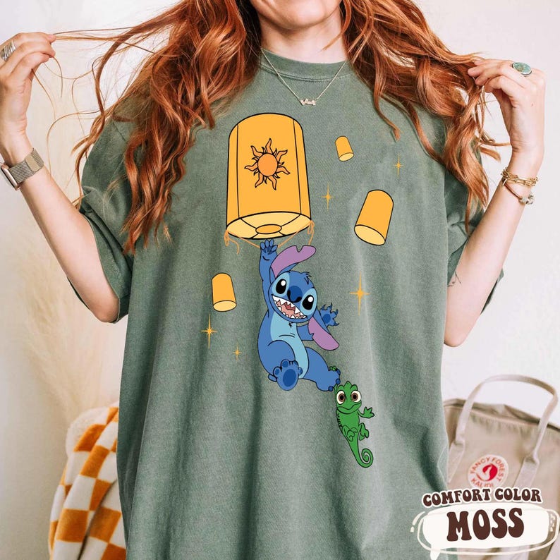 May include: An olive green t-shirt featuring a graphic print of a blue alien character hanging from a large yellow lantern with a sun design. Smaller yellow lanterns and a small green lizard character are also depicted on the shirt fabric.
