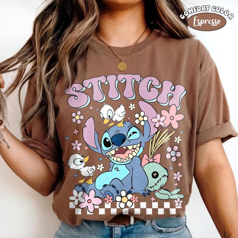 May include: An espresso brown t-shirt featuring a graphic of the blue character Stitch with a small white duck on its head, a green doll, and flowers. The text STITCH is printed in large purple letters above the character, with a checkered pattern below.