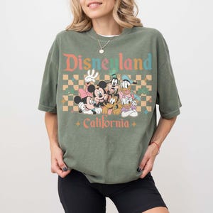 May include: Olive green t-shirt featuring a graphic of Disney characters including Mickey and Minnie Mouse, Goofy, Donald Duck, and Pluto. The text "Disneyland California" is printed above and below the characters in a retro font. The shirt has a relaxed fit.