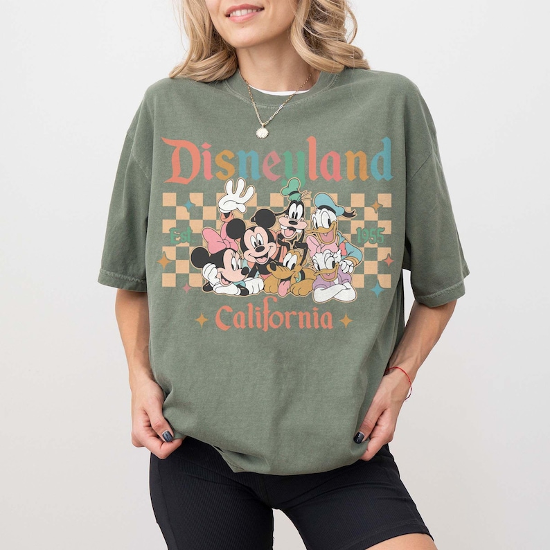 May include: Olive green t-shirt featuring a graphic of Disney characters including Mickey and Minnie Mouse, Goofy, Donald Duck, and Pluto. The text "Disneyland California" is printed above and below the characters in a retro font. The shirt has a relaxed fit.