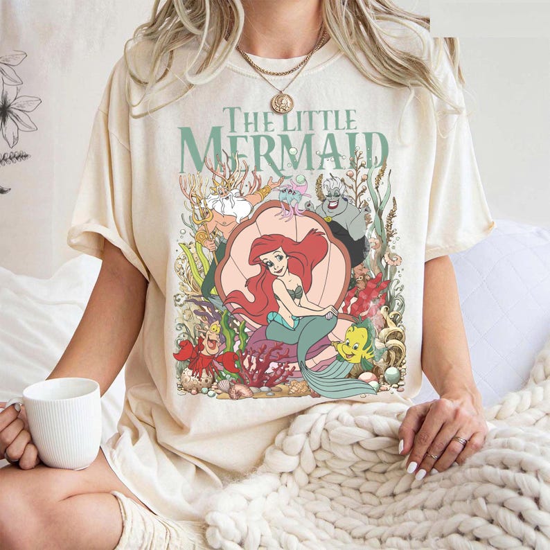 May include: Cream-colored t-shirt featuring a colorful graphic from "The Little Mermaid." The design includes Ariel, Flounder, Sebastian, and other characters. The words "The Little Mermaid" are printed above the characters in a seafoam green color.