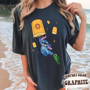 May include: A charcoal gray t-shirt featuring a graphic print of the blue character Stitch holding a yellow floating lantern with a sun design, accompanied by a small green lizard character and several smaller yellow lanterns with star accents.