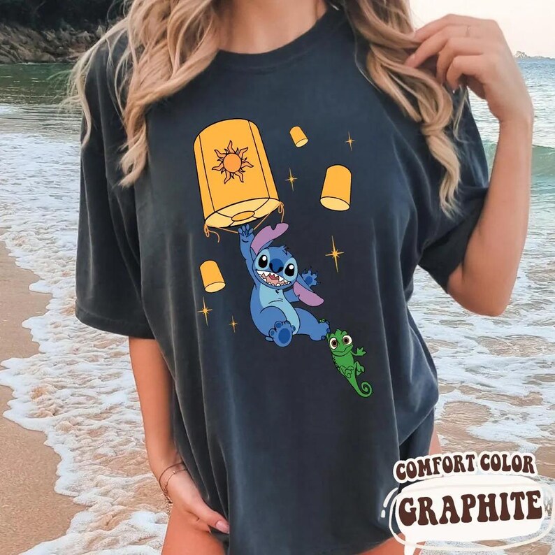 May include: A charcoal gray t-shirt featuring a graphic print of the blue character Stitch holding a yellow floating lantern with a sun design, accompanied by a small green lizard character and several smaller yellow lanterns with star accents.