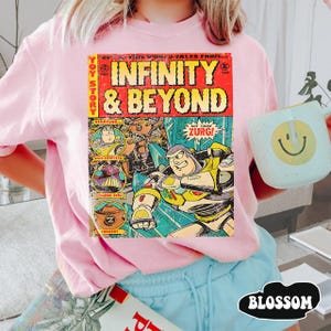 Może przedstawiać: R&oacute;żowa bluza z nadrukiem w stylu komiksowym "INFINITY & BEYOND" i "TOY STORY". Grafika przedstawia Buzza Astrala i Zurga. Widoczny jest r&oacute;wnież jasnoniebieski kubek z uśmiechniętą buzią.