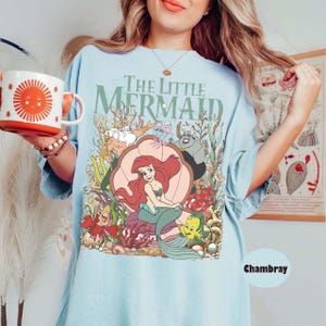 May include: Light blue t-shirt featuring the text "The Little Mermaid" above a colorful graphic of Ariel, Flounder, Sebastian, and other characters. The shirt has a relaxed fit and is made of soft material.