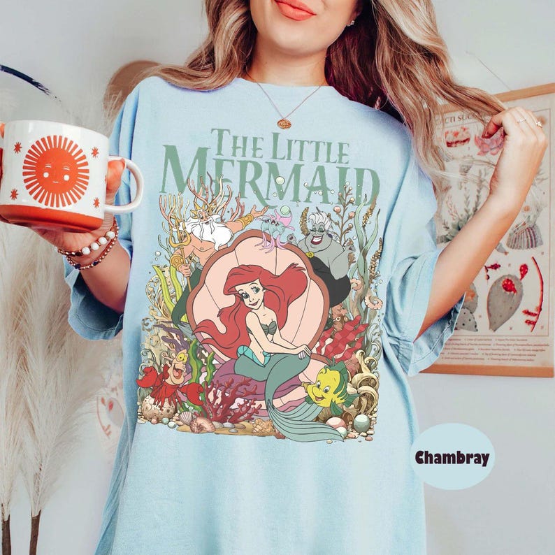 May include: Light blue t-shirt featuring the text "The Little Mermaid" above a colorful graphic of Ariel, Flounder, Sebastian, and other characters. The shirt has a relaxed fit and is made of soft material.