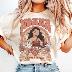 Op de afbeelding: Cr&egrave;mekleurig T-shirt met een afbeelding van Moana, een Disney-prinses, en het woord "Moana" erboven. Het ontwerp bevat vlinders, bloemen en de tekst "EST 2016 DISNEY PRINCESS".
