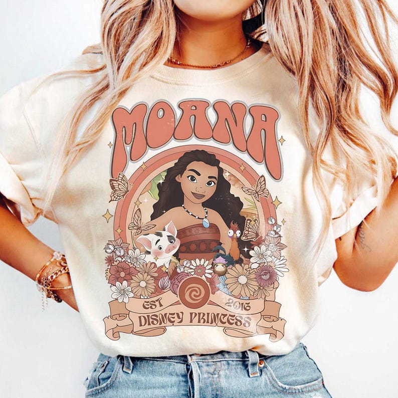 Op de afbeelding: Cr&egrave;mekleurig T-shirt met een afbeelding van Moana, een Disney-prinses, en het woord "Moana" erboven. Het ontwerp bevat vlinders, bloemen en de tekst "EST 2016 DISNEY PRINCESS".