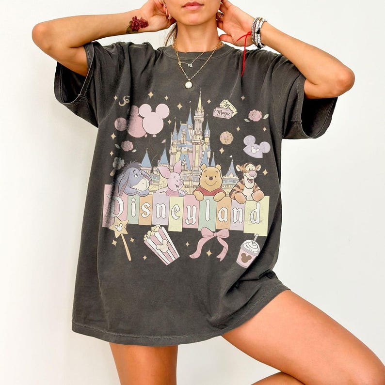 May include: Dark grey t-shirt with a Disneyland graphic. The design features a castle, Winnie the Pooh characters, and the Disneyland logo in pastel colours. Additional elements include popcorn, a bow, and a Mickey Mouse balloon.