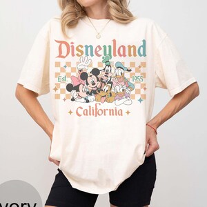 May include: Ivory-colored t-shirt featuring a retro Disneyland graphic with Mickey Mouse, Minnie Mouse, Goofy, Donald Duck, and Pluto. The design includes the text "Disneyland Est. 1955 California" in a colorful, vintage font. The shirt has short sleeves.