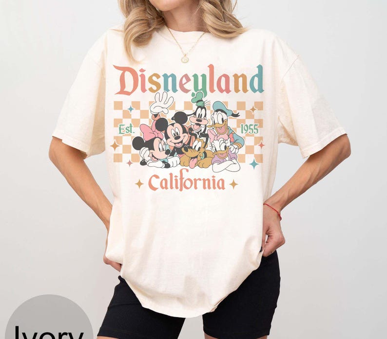 May include: Ivory-colored t-shirt featuring a retro Disneyland graphic with Mickey Mouse, Minnie Mouse, Goofy, Donald Duck, and Pluto. The design includes the text "Disneyland Est. 1955 California" in a colorful, vintage font. The shirt has short sleeves.