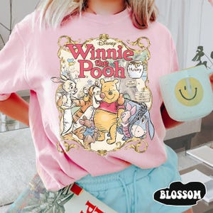 May include: Pink t-shirt featuring a graphic of Winnie the Pooh and friends. The design includes the text "Winnie the Pooh" in a decorative font, along with illustrations of Pooh, Piglet, Tigger, Eeyore, and Rabbit. The shirt is a light pink color.