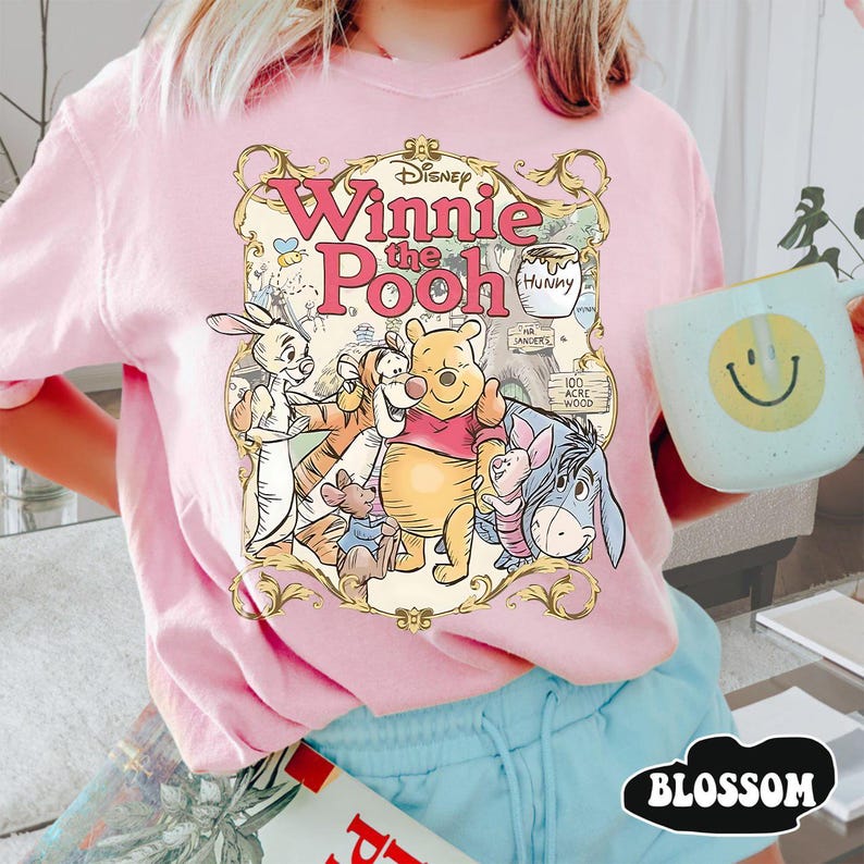 May include: Pink t-shirt featuring a graphic of Winnie the Pooh and friends. The design includes the text "Winnie the Pooh" in a decorative font, along with illustrations of Pooh, Piglet, Tigger, Eeyore, and Rabbit. The shirt is a light pink color.
