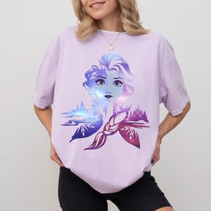 以下が含まれることがあります： 女性の顔のグラフィックデザインが特徴の薄紫色のTシャツ。青と紫の色合いが使われています。デザインには城と風景の要素が含まれています。Tシャツはカジュアルなスタイルです。