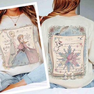 Pode incluir: Camisola creme com uma ilustra&ccedil;&atilde;o colorida de Elsa e Anna de Frozen na frente. A parte de tr&aacute;s da camisola mostra Olaf, flocos de neve e partituras com o texto "Let It Go". Acentos florais emolduram o design.