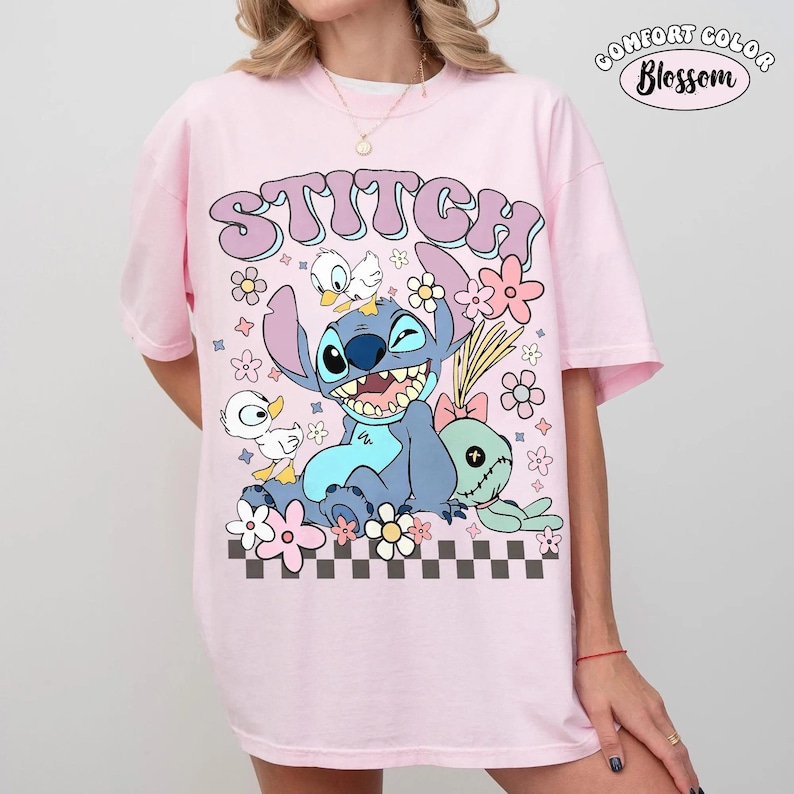 May include: A light pink t-shirt featuring a graphic print of the character Stitch, two small white ducks, and a green doll. The text "STITCH" appears in large purple letters above the characters, surrounded by pink and yellow flowers and a checkered pattern.