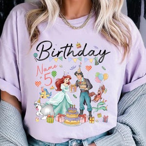 Pode incluir: Uma camiseta lil&aacute;s com um gr&aacute;fico colorido com a palavra "Birthday" e o n&uacute;mero "5". O design incorpora personagens de desenhos animados, um bolo e bal&otilde;es, criando um tema de anivers&aacute;rio festivo.