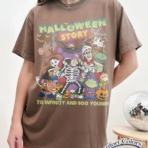 May include: Espresso-coloured t-shirt with a Halloween-themed graphic featuring cartoon characters and the text "HALLOWEEN STORY" and "TO INFINITY AND BOO YOND!". The shirt is a Comfort Colors brand.