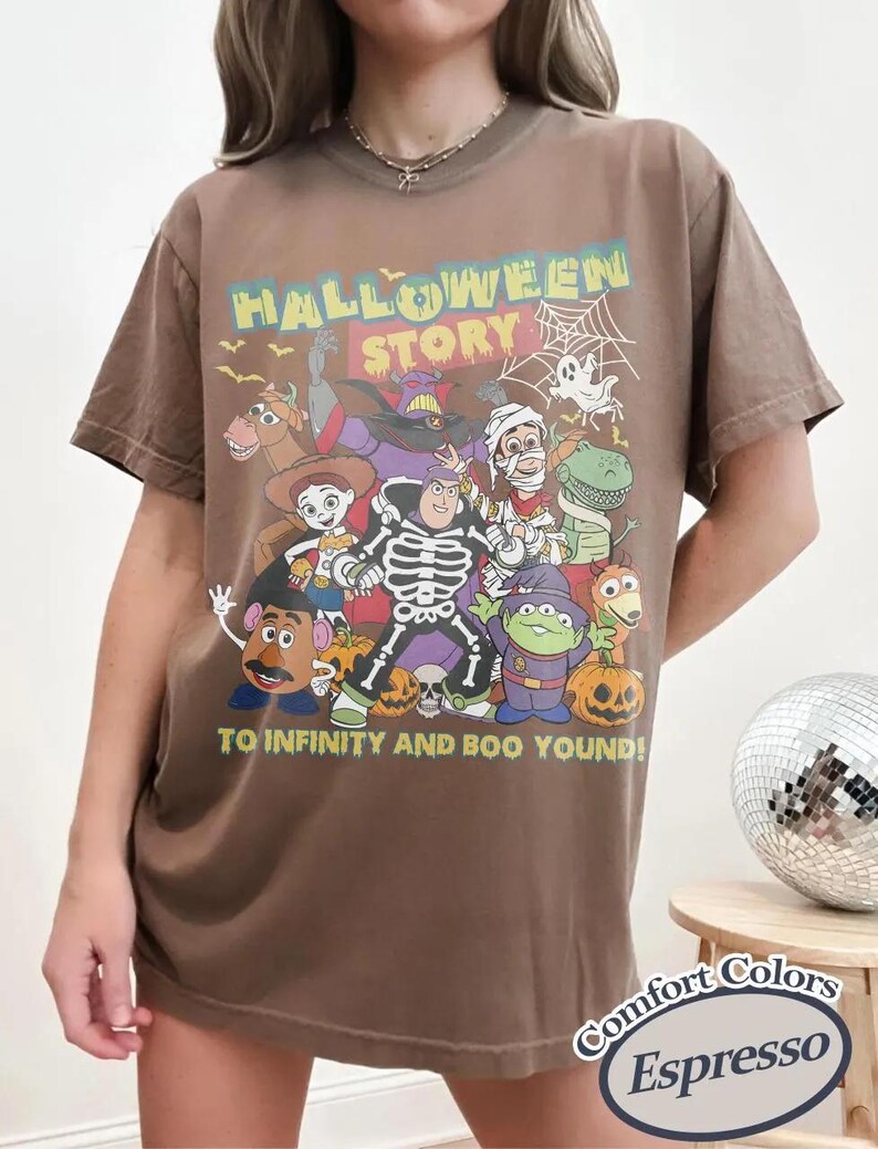 May include: Espresso-coloured t-shirt with a Halloween-themed graphic featuring cartoon characters and the text "HALLOWEEN STORY" and "TO INFINITY AND BOO YOND!". The shirt is a Comfort Colors brand.