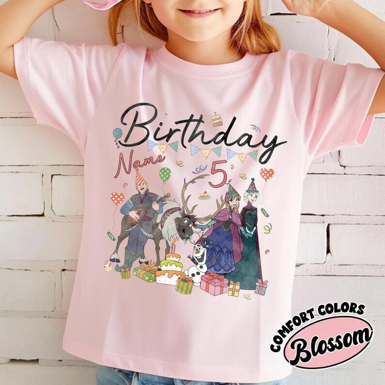May include: A light pink t-shirt with a colorful graphic featuring the text "Birthday" and "Name" above the number "5". The graphic includes cartoon characters, a cake, and presents. The shirt has the words "Comfort Colors" and "Blossom" in a circular design.