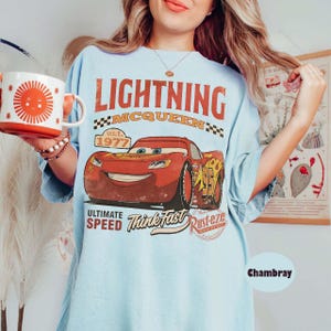 Puede incluir: Camiseta azul claro con un gr&aacute;fico de Rayo McQueen de la pel&iacute;cula Cars. El dise&ntilde;o incluye el texto "LIGHTNING MCQUEEN" y "ULTIMATE SPEED". En la imagen se sostiene una taza roja y blanca con un dise&ntilde;o de sol.