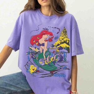 K&ouml;nnte beinhalten: Ein lila T-Shirt mit einer farbenfrohen Grafik von Arielle, der Meerjungfrau. Die Illustration zeigt Arielle mit roten Haaren, einem gr&uuml;nen Schwanz und verschiedenen Meereskreaturen. Der Text "THE LITTLE MERMAID" befindet sich unten.