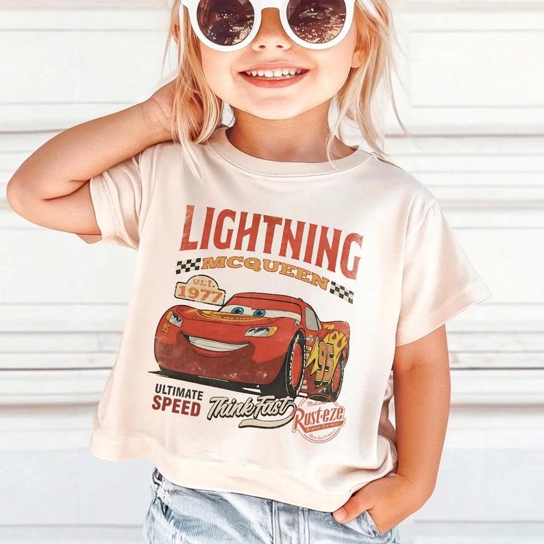 Puede incluir: Camiseta beige claro con un dise&ntilde;o de Lightning McQueen, un coche de carreras rojo. El texto "LIGHTNING MCQUEEN" y "est. 1977" son en rojo. El texto adicional incluye "ULTIMATE SPEED Think Fast Rust-eze".