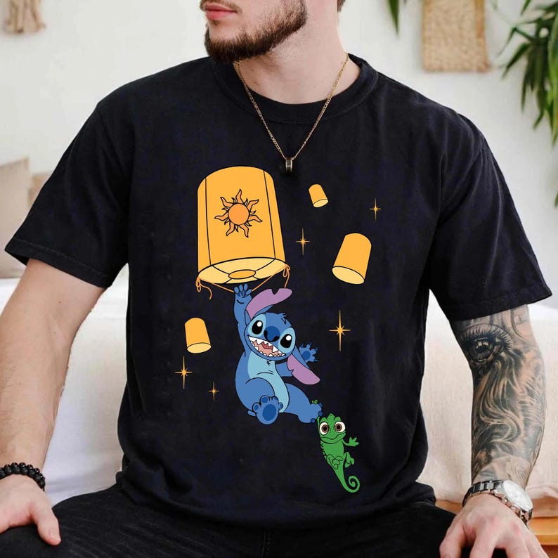 May include: A black short-sleeved t-shirt featuring a graphic print of a blue alien character holding onto a large yellow lantern with a sun design. Several smaller yellow lanterns and four-pointed star shapes are scattered around the character.