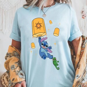 May include: A light blue t-shirt featuring a graphic print of a blue alien character holding onto a large yellow lantern with a sun symbol, accompanied by a small green lizard character and several smaller floating yellow lanterns and stars.