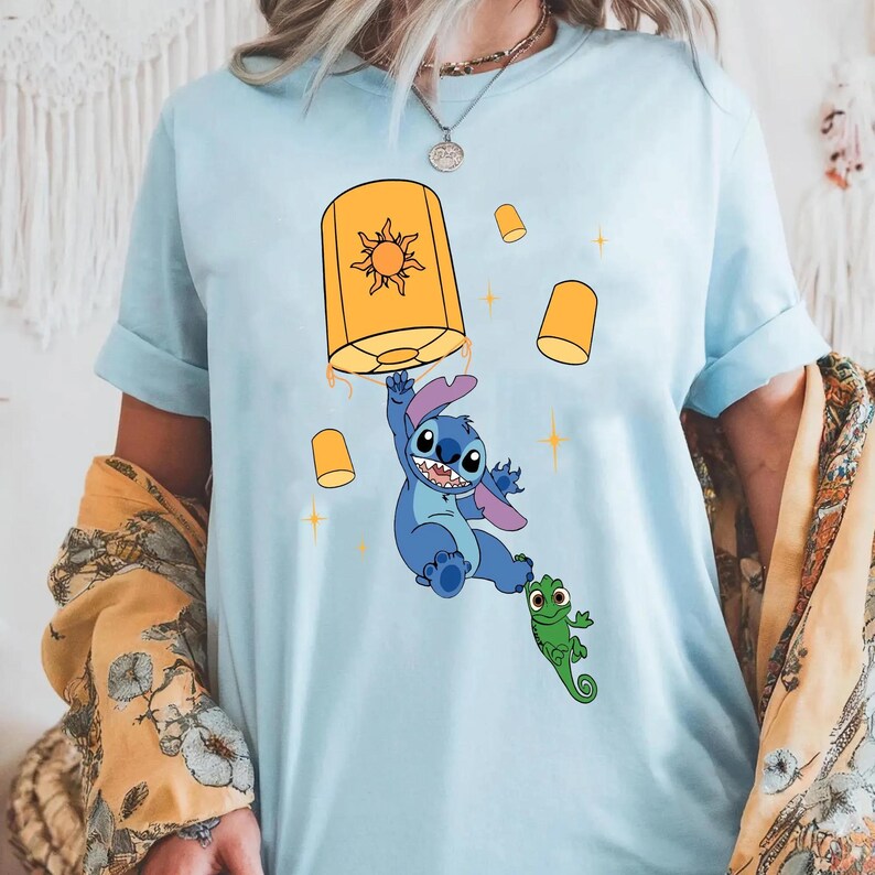 May include: A light blue t-shirt featuring a graphic print of a blue alien character holding onto a large yellow lantern with a sun symbol, accompanied by a small green lizard character and several smaller floating yellow lanterns and stars.