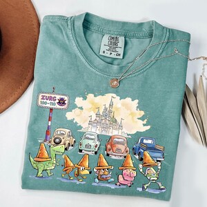 May include: A teal Comfort Colors t-shirt featuring a whimsical design with cartoon characters, cars, and a castle. The shirt includes the text "Zurg 100-110" on a road sign. A silver necklace with a pendant is also visible.