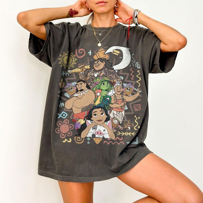 May include: A charcoal gray short-sleeve t-shirt featuring a graphic print of characters from the movie Moana, including Moana, Maui, Pua, Heihei, and Gramma Tala, surrounded by colorful tribal-inspired patterns and symbols on the front.