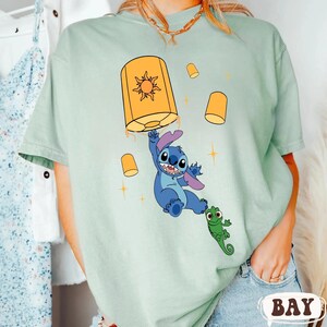 May include: A sage green t-shirt featuring a graphic of a blue alien character holding onto a large yellow lantern with a sun symbol, accompanied by a small green chameleon character and several smaller floating yellow lanterns with star accents.