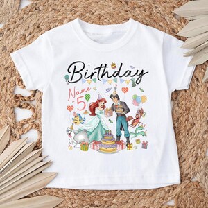 Pode incluir: T-shirt branco de manga curta com um gr&aacute;fico colorido de anivers&aacute;rio. O texto "Birthday" est&aacute; em preto, o n&uacute;mero "5" em rosa, com personagens de desenhos animados, um bolo e bal&otilde;es.