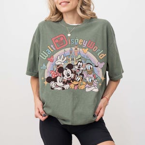 May include: Olive green t-shirt featuring a colorful graphic of Disney characters, including Mickey and Minnie Mouse, Donald Duck, and Goofy, with the text "Walt Disney World" above a rainbow and castle.