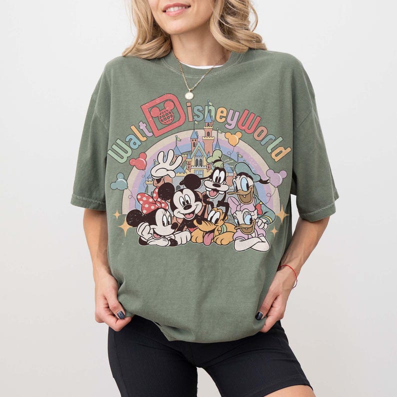 May include: Olive green t-shirt featuring a colorful graphic of Disney characters, including Mickey and Minnie Mouse, Donald Duck, and Goofy, with the text "Walt Disney World" above a rainbow and castle.