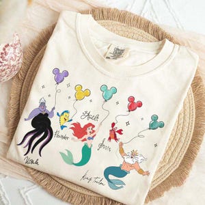 Puede incluir: Camiseta color crema con los personajes de La Sirenita de Disney: &Uacute;rsula, Flounder, Ariel, Sebasti&aacute;n y el Rey Trit&oacute;n. Cada personaje sostiene un globo con forma de Mickey Mouse. La camiseta tiene las palabras "&Uacute;rsula", "Flounder", "Ariel", "Sebasti&aacute;n" y "Rey Trit&oacute;n".