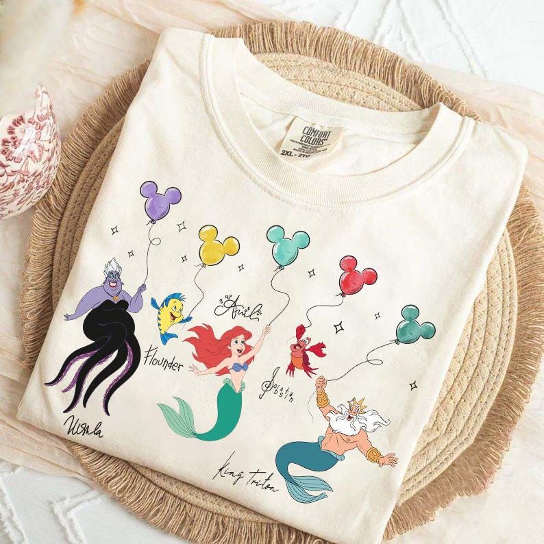 Puede incluir: Camiseta color crema con los personajes de La Sirenita de Disney: &Uacute;rsula, Flounder, Ariel, Sebasti&aacute;n y el Rey Trit&oacute;n. Cada personaje sostiene un globo con forma de Mickey Mouse. La camiseta tiene las palabras "&Uacute;rsula", "Flounder", "Ariel", "Sebasti&aacute;n" y "Rey Trit&oacute;n".