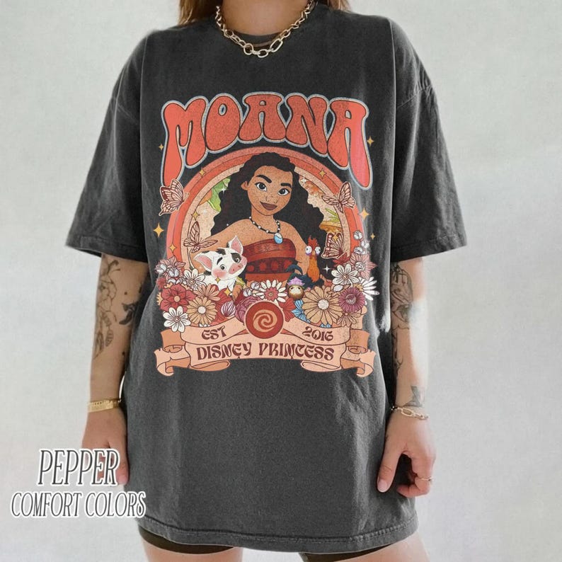 Op de afbeelding: Donkergrijs T-shirt met een kleurrijke afbeelding van een Disney-prinses. Het ontwerp bevat het woord "Moana" in oranje, een portret van de prinses en de tekst "Est Disney Princess 2016". Het shirt heeft ook de tekst "Pepper Comfort Colors".