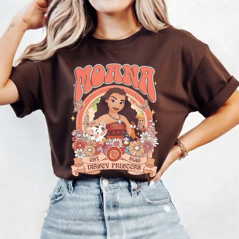 Op de afbeelding: Bruin t-shirt met een retro-ontwerp van Moana, omringd door bloemen en vlinders. Het ontwerp bevat het woord "Moana" in een vintage lettertype en de tekst "Est 2016 Disney Princess" op een banner.