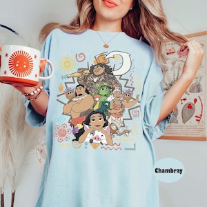 May include: A light blue t-shirt featuring a colorful graphic print of characters from the movie Moana, including Moana, Maui, Pua, Heihei, and Gramma Tala. The shirt is paired with a white mug with a large orange sun design on the front.